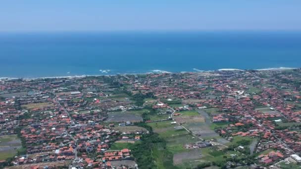 Drone voar de Canggu de cima em tempo ensolarado Indonésia Bali, telhados vermelhos visíveis, campos de arroz verde e do oceano no fundo — Vídeo de Stock