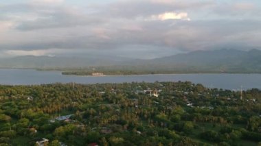 Palmiye ağaçlarının tepesinden havalan ve adayı ve anakara dağlarını ayıran bir cami ve su görülebilir durumda.