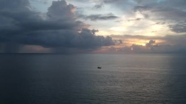 İnsansız hava aracı gün batımında okyanusun üzerinde ilerler. Merkezde turuncu-mavi renkli bulutlar içinde bir tekne görülebilir.