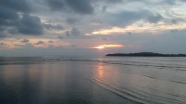 Kamera okyanusun üzerinde, bulutlu bir gökyüzüne karşı gün batımına doğru zum yapıyor ve pembe-turuncu bir güneş halkası görülebiliyor.