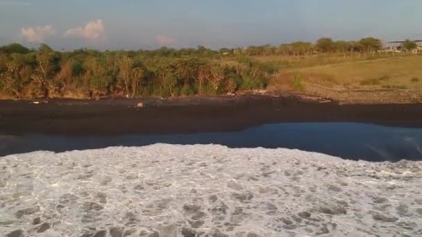 Камера летить далеко від пляжу чорного піску на заході сонця, видно величезні білі хвилі та піну — стокове відео