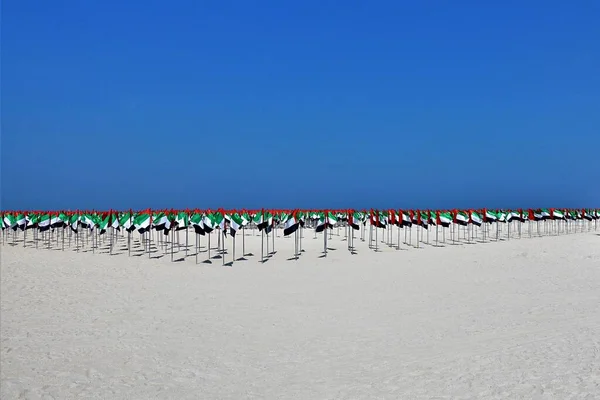 Installation in honor of the celebration of the UAE flag day. Many flags of red, green, white and black colors form a composition on clean light sand under a bright blue sky. They symbolize Arab unity.