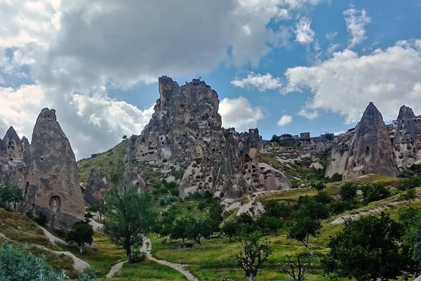 The ancient fortress of Uchisar in Cappadocia. The fortress is carved into the rock. Paths lead through the green lawn. Around peaked tuff rocks. There are clouds in the sky.