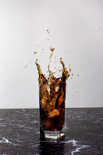 The drink is splashed out of a glass cup, spatter and a splash of beverage. Throwing ice in a glass with a sweet brown drink and a lot of spray.