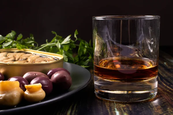A glass of whiskey and an elite appetizer on a black background. Olives, pickled mushrooms, champignons, sprats and an expensive alcoholic drink. — Stock Image Glass Whiskey Elite Appetizer Black Background Olives Pickled Mushrooms Champignons Royalty Free Stock Images