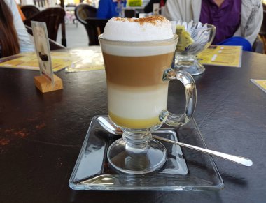 Bir fincan vanilyalı cappuccino içiyorum, cam tabakta, kafede.