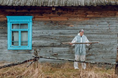 Beyaz koruyucu takım elbiseli bir adam, köyde eski bir evin yanında tulum giyiyor. Virüse karşı koruyucu giysi.