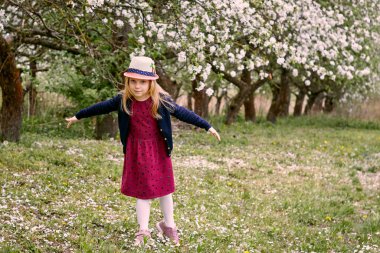 Elma bahçesinde şapkalı ve kırmızı elbiseli bir kız. Yeşil çimenlerin ve çiçek açan ağaçların arka planında.