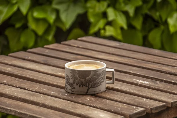 White matte cup with a monochrome pattern and with coffee on a wooden table with a blurred background of green leaves