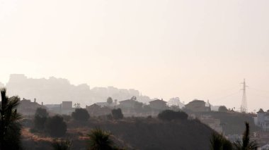 Ev şeklinde bir köy ve ön planda çalılarla birlikte gün doğarken hafif bir sis altında yüksek voltaj direği.