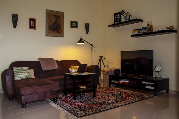 Oturma odası halı, kanepe, dizüstü bilgisayarla aydınlatılmış masa lamba, televizyon standı ve TV, duvarlarda çerçeveli üç poster ve çeşitli objelerle dolu iki raf..