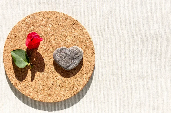 Red flower and a stone in the form of a heart are on a round plate made of cork on a textural background with a sharp shadow from the sun