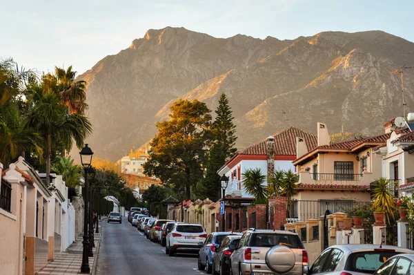 Marbella şehrinde gün batımında Sierra Blanca dağının arka planına karşı park edilmiş arabaları ve özel evleri olan bir cadde..