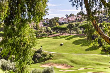 Golf sahasının panoramik manzarası, oyuncuların arka plandaki özel evlere ve ön plandaki ağaçların arasından geçen bulutlu mavi gökyüzüne karşı.