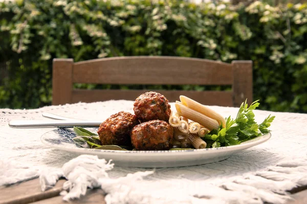 Üzerinde üç köfte, makarna ve maydanoz olan bir tabak bulunan keten masa örtüsü olan ahşap bir masa. Sandalyenin ve bahçe çitlerinin arkasında.