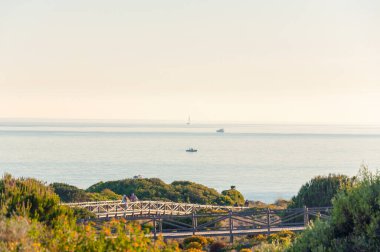 Tıbbi maskeli iki kişi, gün batımında bir yat, tekne ve balıkçılarla dolu bir tekneyle, denizin zeminine karşı ahşap kaldırımlar ve yeşil çalılarla parkta yürüyor..