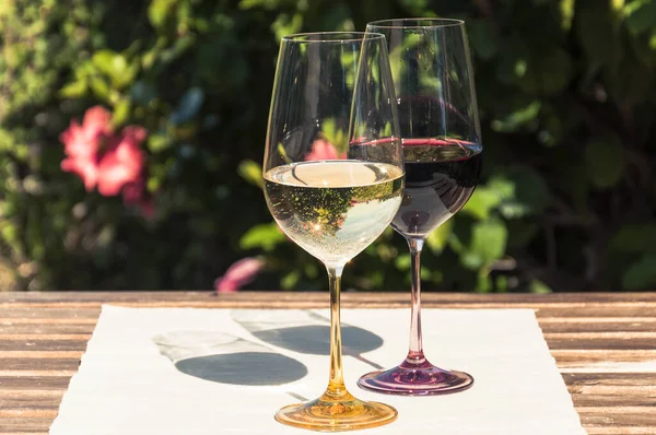 A glass of white and red wine stand on a linen tablecloth on a wooden table on a blurred background of a green bush with red flowers