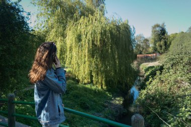 Mavi ceketli ve kot pantolonlu bir bayan bahar parkında, dere köprüsünün üzerinde telefonla konuşuyor ve arka planda ağlayan bir söğüt ağacı var.