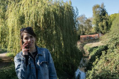 Mavi ceketli ve kot pantolonlu bir kadın bir dere köprüsünün üzerindeki parkta telefonla konuşuyor ve arka planda ağlayan bir söğüt ağacı var.