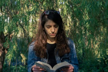 Mavi ceketli kadın söğüt ağacının altında roman okuyor.