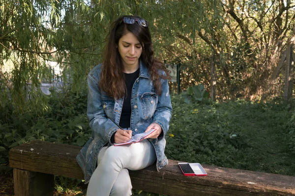 Lady drawing on a red notebook hanged on her knees while is sitting on a wooden bench