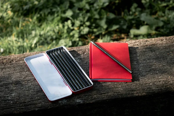 red notebook and a set of drawing pencils hanged on a outdoor wooden bench