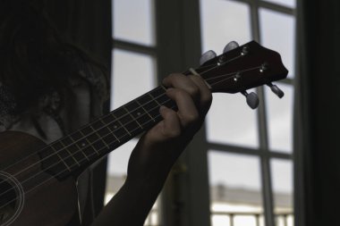 Ukulele 'nin boynuna yakın plan çekimde arka planda parlak pencereli bir kız oynuyor.