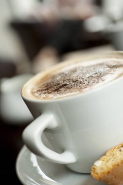 Köpüğünde çikolata tozu olan bir Cappuccino. Kafe tarzı bir tabağın kenarına İtalyan bisküvisi konmuş.