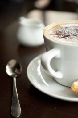Köpüğünde çikolata tozu olan bir Cappuccino. Kafe tarzı bir tabağın kenarına İtalyan bisküvisi konmuş.