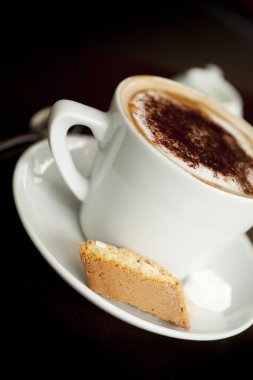 Köpüğünde çikolata tozu olan bir Cappuccino. Kafe tarzı bir tabağın kenarına İtalyan bisküvisi konmuş.