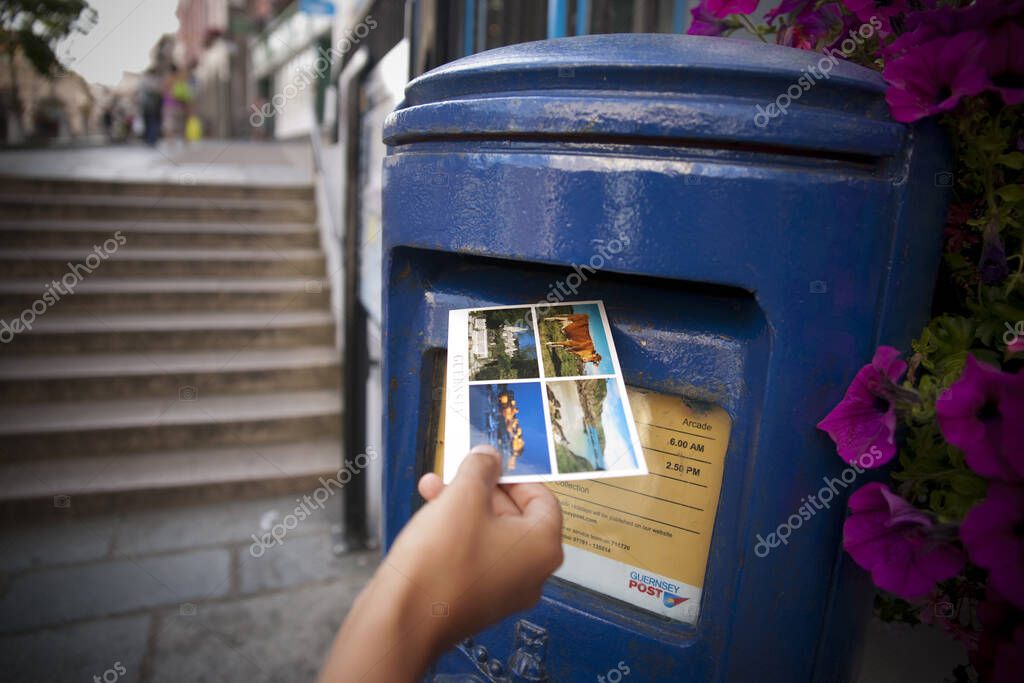 A Hand putting a postcard in a Blue Guernsey Post Box unique to Guernsey in the town of St Pierre Port (St Peter Port), the main settlement of Guernsey, The Channel Islands, UK - 11th July 2013