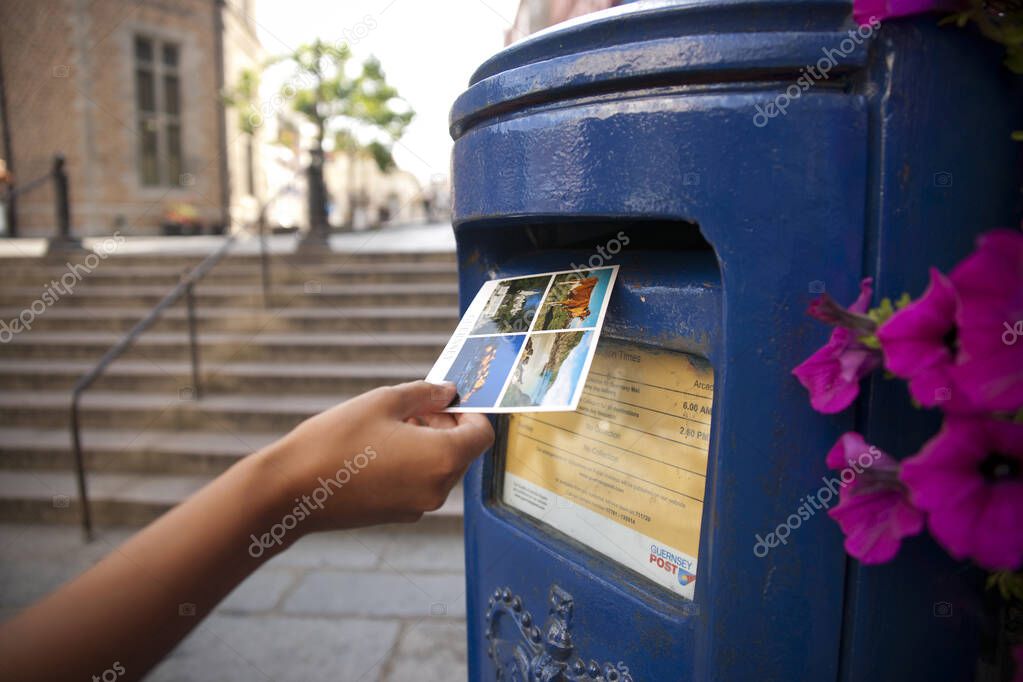 A Hand putting a postcard in a Blue Guernsey Post Box unique to Guernsey in the town of St Pierre Port (St Peter Port), the main settlement of Guernsey, The Channel Islands, UK - 11th July 2013
