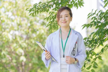 Açık havada gülümseyen beyaz önlüklü bir kadın doktor portresi.