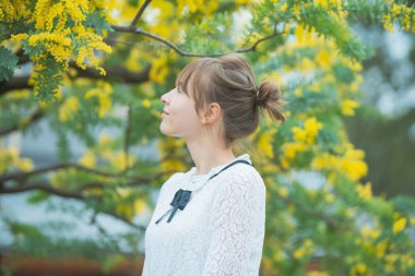 Beyaz tenli genç bir kadın çiçek açan mimoza çiçeklerine bakıyor.