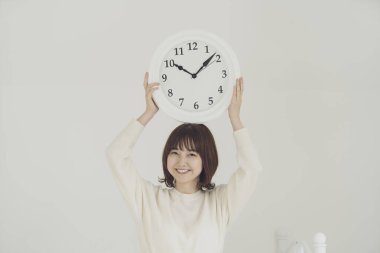 Gülümseyen Asyalı genç kadın kafasında beyaz duvar saati tutuyor.