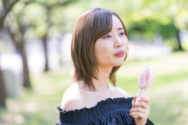 Genç Asyalı kadın yüzünün yanında taşınabilir bir vantilatör tutuyor.