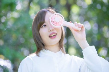 Genç Asyalı kadın yüzünün yanında taşınabilir bir vantilatör tutuyor.