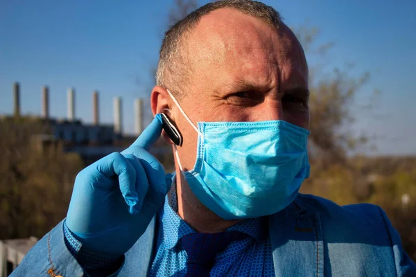 Coronavirus. Quarantine. Mature man in a medical mask and gloves. Industrial landscape. Bad ecology. Business man on the territory of the enterprise. Speaks on a smartphone using a headset.