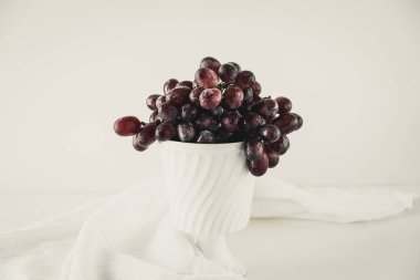 Beyaz bir masanın üzerinde duran eski beyaz bir porselen kasedeki koyu kırmızı üzümler..