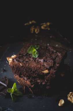 İki parça cevizli kek, koyu çikolata, tuz ve nane yaprakları bir tabakta ve koyu kahverengi arka planda. Dikey.