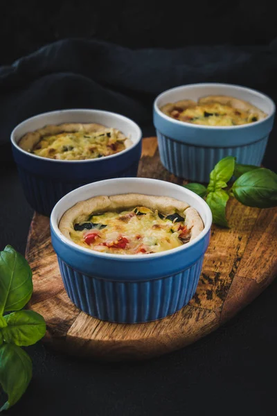 Baked vegetable tartlets in blue colored tart dishes on a wooden board on dark blue background. Vertical closeup.