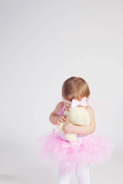 A little girl in a full skirt and with wings on her back hugs a bear on a white background