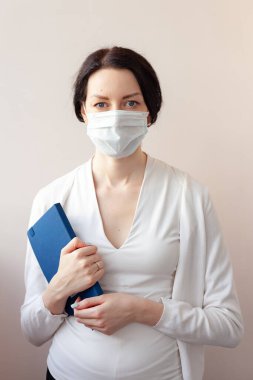Koruyucu maskeli bir kadın beyaz bir ceketle arka planda duruyor ve elinde bir günlük tutuyor. Not alan bir tıp uzmanı.