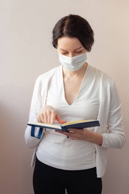 Koruyucu maskeli bir kadın beyaz bir ceketle arka planda duruyor ve elinde bir günlük tutuyor. Not alan bir tıp uzmanı.