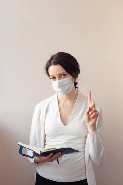Koruyucu maskeli bir kadın beyaz bir ceketle arka planda duruyor ve elinde bir günlük tutuyor. Not alan bir tıp uzmanı.