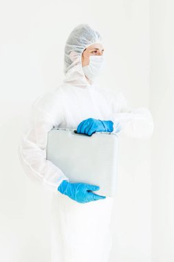 Koruyucu takım elbiseli, eldivenli ve maskeli bir adam elinde bir bavulla elinde testler için yürüyor. Koronavirüsle mücadele kavramı. Beyaz bir arkaplanda izole edilmiş