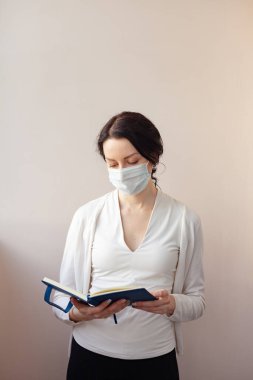 Koruyucu maskeli bir kadın beyaz bir ceketle arka planda duruyor ve elinde bir günlük tutuyor. Not alan bir tıp uzmanı.
