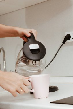 Kız elektrikli çaydanlıktan bardağa su döküyor. Beyaz bluzlu genç bir kadın mutfakta duruyor..