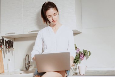 Kız mutfak masasında oturan bir dizüstü bilgisayarla çalışıyor. Mutfakta beyaz gömlekli ve kot şortlu genç bir kadın var. Karantinada evde çalışma kavramı