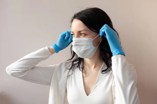 A woman in gloves and a protective mask stands on a light background in a white jacket. The concept of suppression of coronavirus